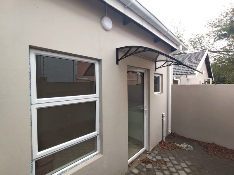 A view of the rear exterior of the flat, featuring a paved courtyard, modern aluminium window frames, a glass door, and a protective awning.