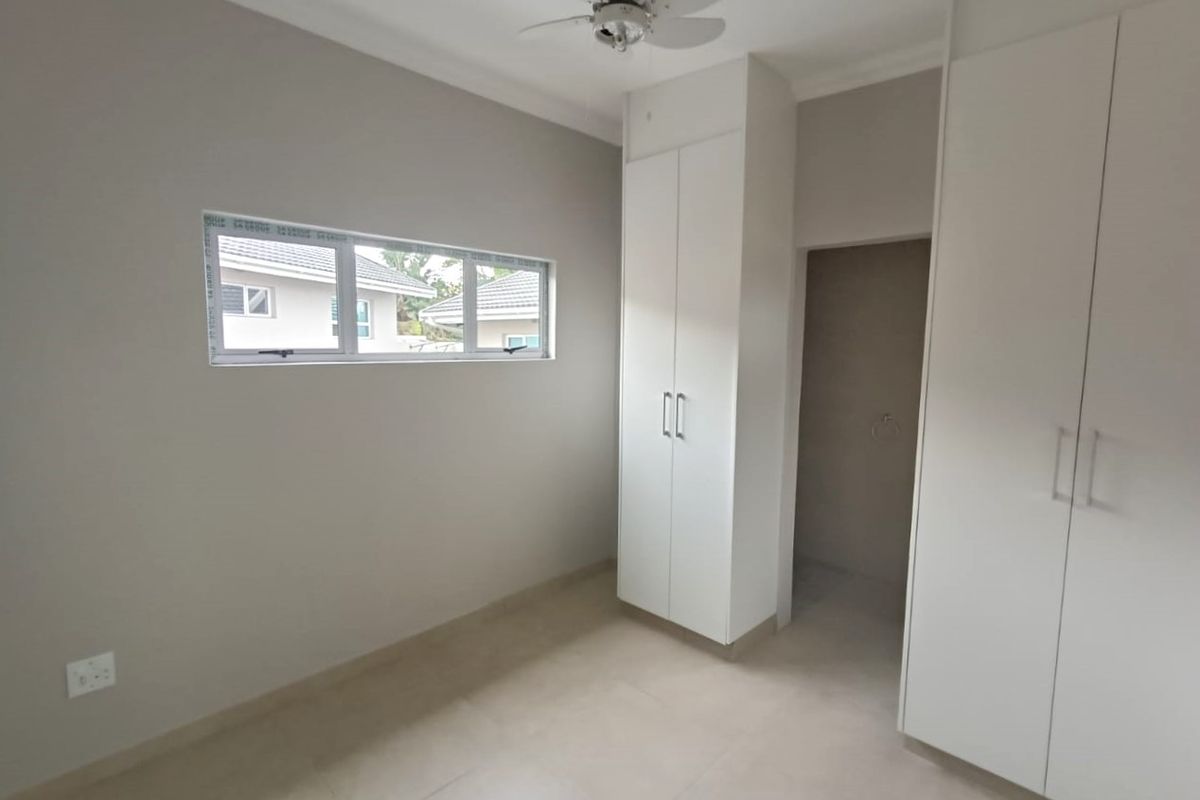 A clean, empty bedroom featuring neutral tiled flooring, built-in white cupboards, a ceiling fan, and an aluminium-framed window.