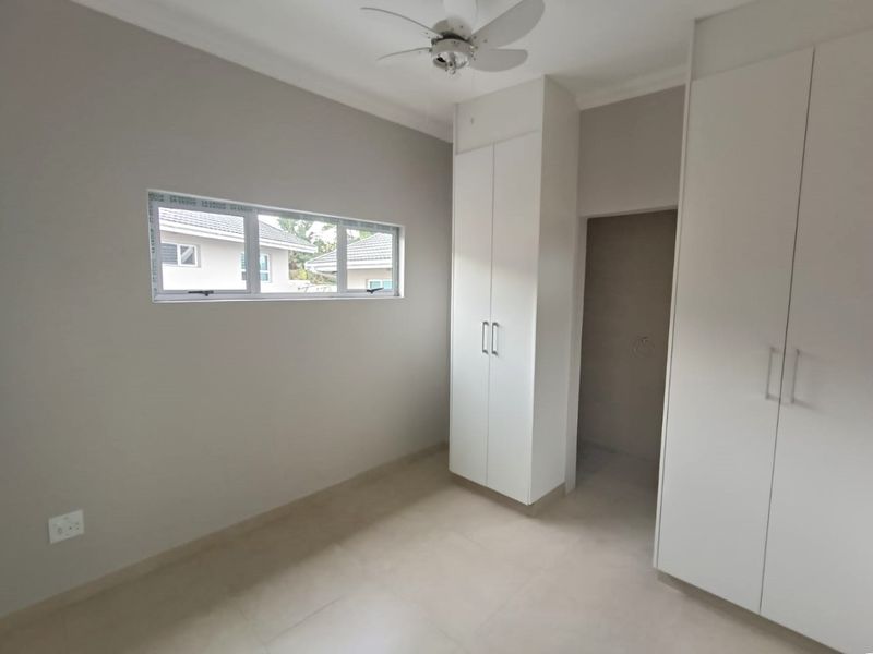A clean, empty bedroom featuring neutral tiled flooring, built-in white cupboards, a ceiling fan, and an aluminium-framed window.