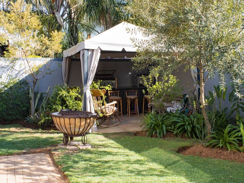A covered outdoor gazebo area featuring a built-in bar counter, set within a landscaped garden with paved flooring.