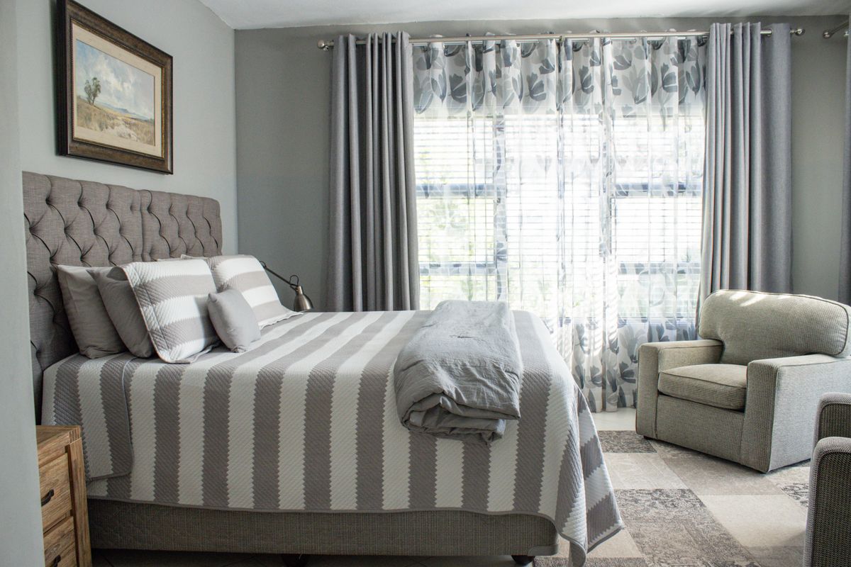 A well-maintained bedroom featuring neutral wall tones, large windows with fitted blinds, and layered curtain treatments.