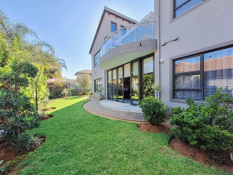 Rear view of the property featuring a landscaped garden, paved patio area, and a curved balcony with a modern glass balustrade.