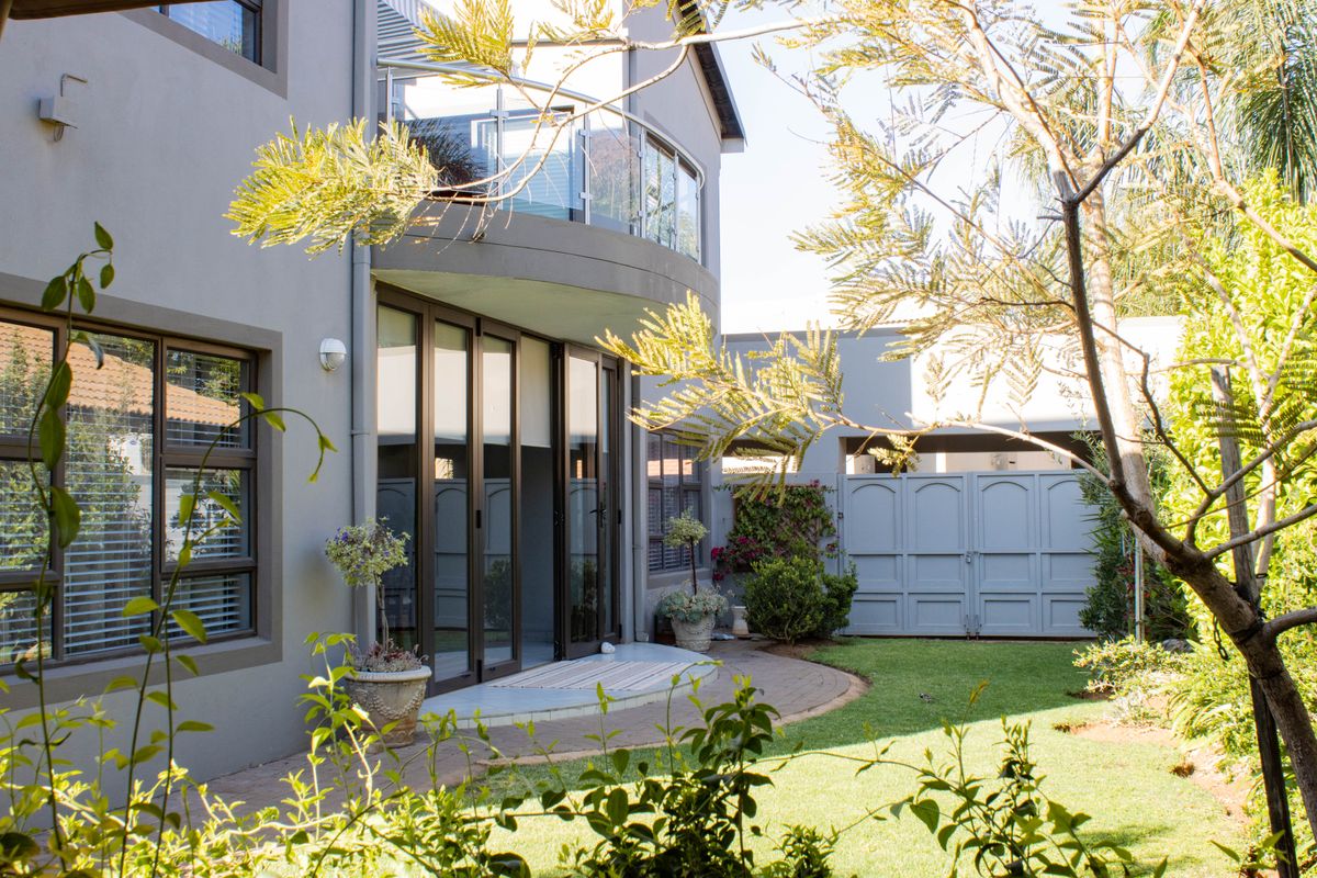 Rear view of a modern two-story house featuring a curved balcony with glass balustrades, paved patio area, and a well-maintained garden with gated access.