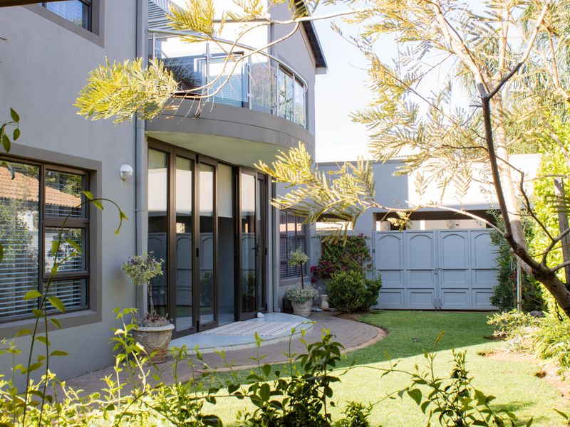 Rear view of a modern two-story house featuring a curved balcony with glass balustrades, paved patio area, and a well-maintained garden with gated access.