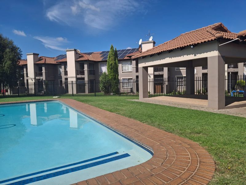 Communal pool area featuring a swimming pool with brick coping, a covered patio structure, and well-maintained lawn within a secure complex.