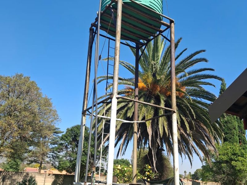 An elevated green water storage tank mounted on a metal support structure located within a residential garden setting.