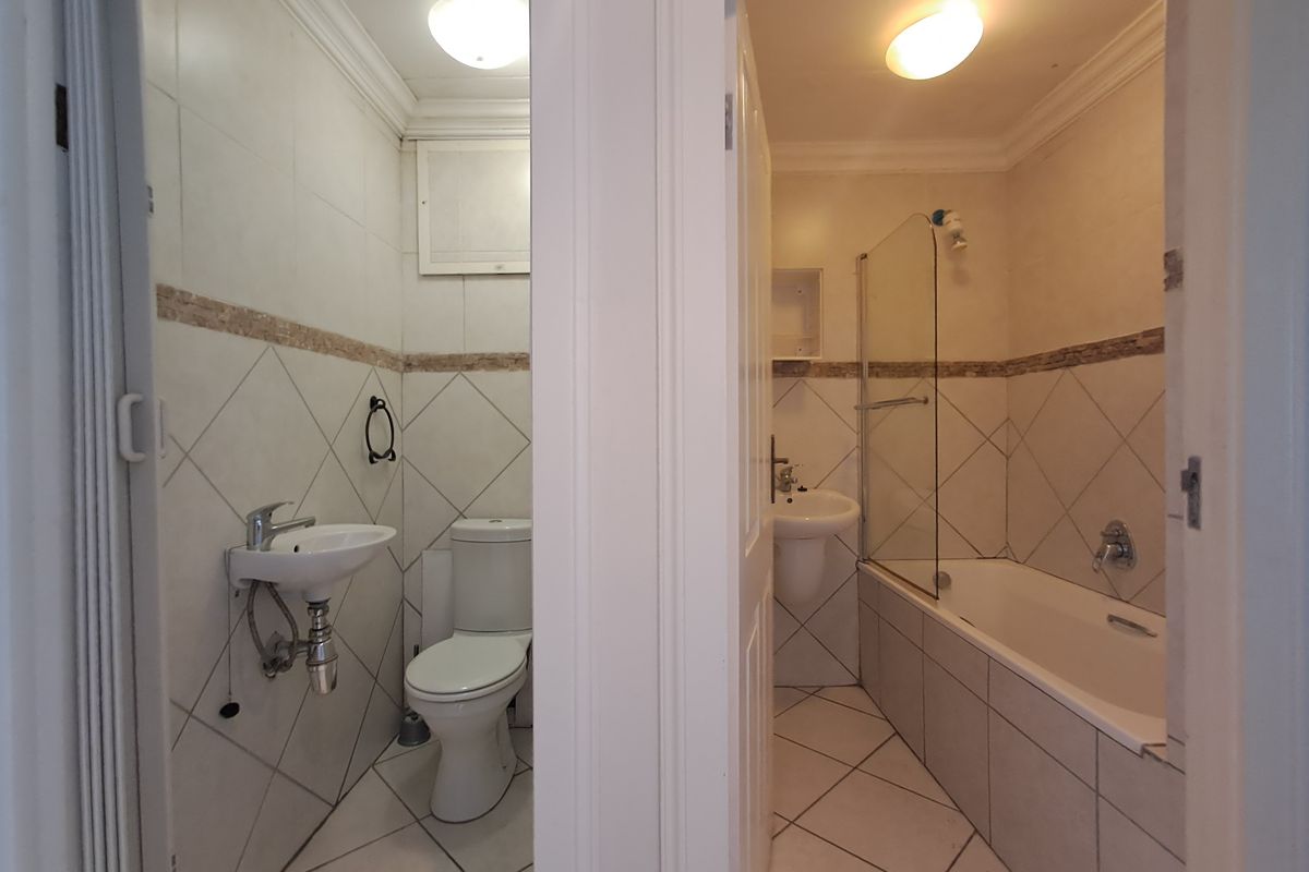 A split-view bathroom featuring a separate toilet area with a basin and a main bathroom area equipped with a bathtub, shower screen, and pedestal basin.