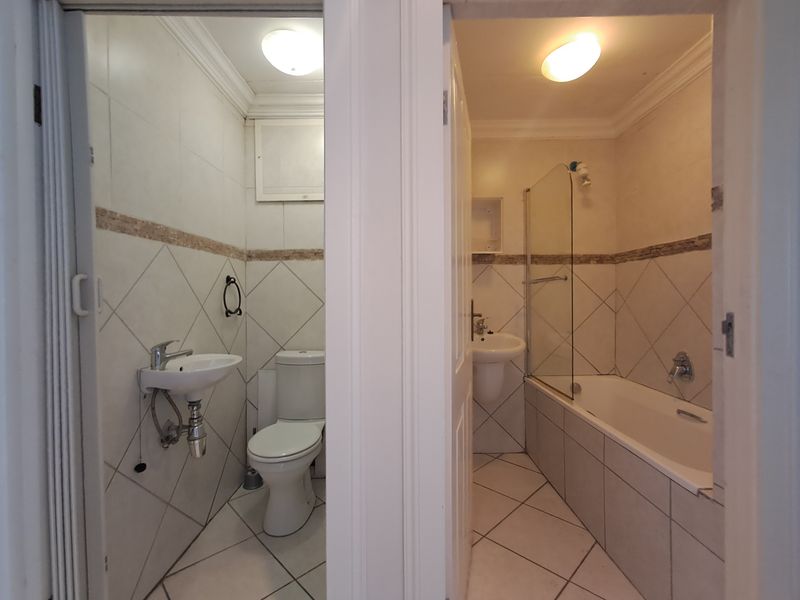 A split-view bathroom featuring a separate toilet area with a basin and a main bathroom area equipped with a bathtub, shower screen, and pedestal basin.