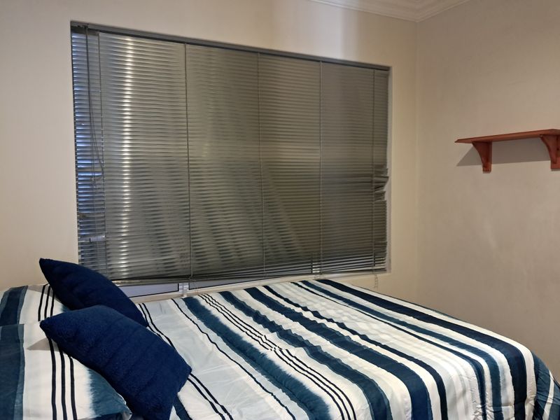 A bedroom featuring a window with venetian blinds and a wall-mounted shelf, providing a functional space with neutral wall finishes.
