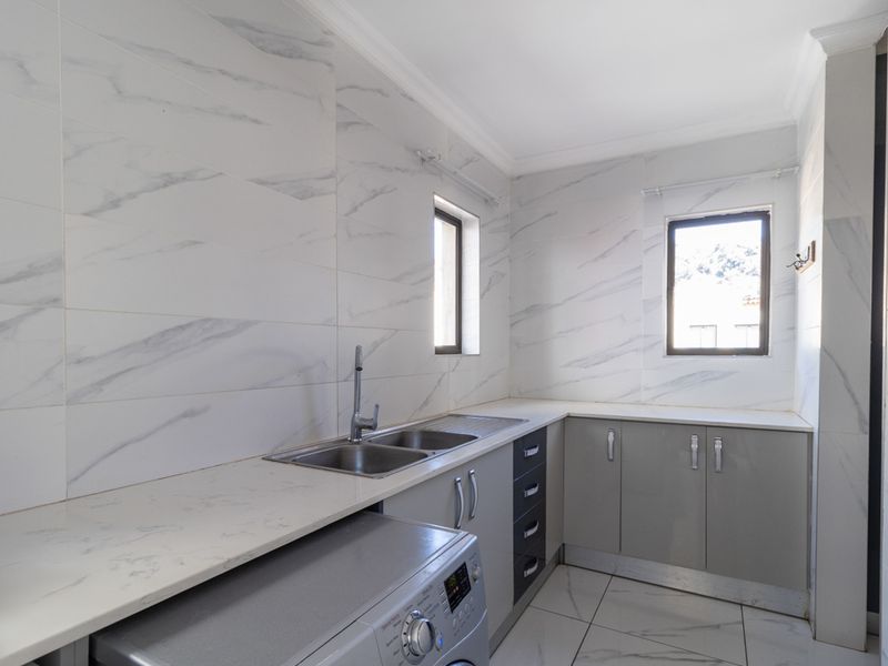 A clean, modern scullery featuring marble-look wall and floor tiles, ample built-in cabinetry, and a double stainless steel sink with a chrome mixer tap.