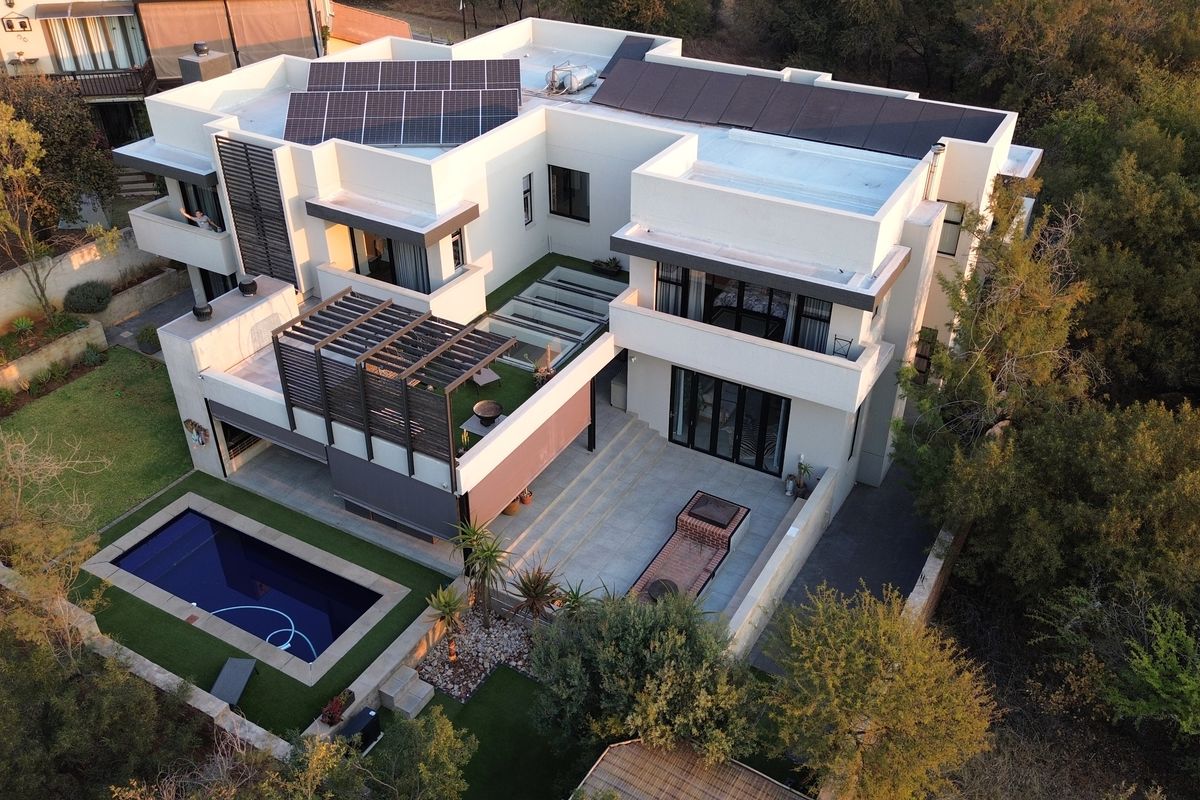 Aerial view of a modern multi-level residence featuring a swimming pool, spacious tiled patio with built-in braai, pergola, and rooftop solar panel installation.