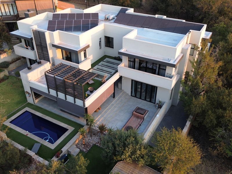 Aerial view of a modern multi-level residence featuring a swimming pool, spacious tiled patio with built-in braai, pergola, and rooftop solar panel installation.