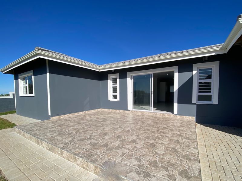 A modern, tiled outdoor patio area featuring a sliding glass door entrance, clean grey exterior walls, and paved surroundings under a clear blue sky.