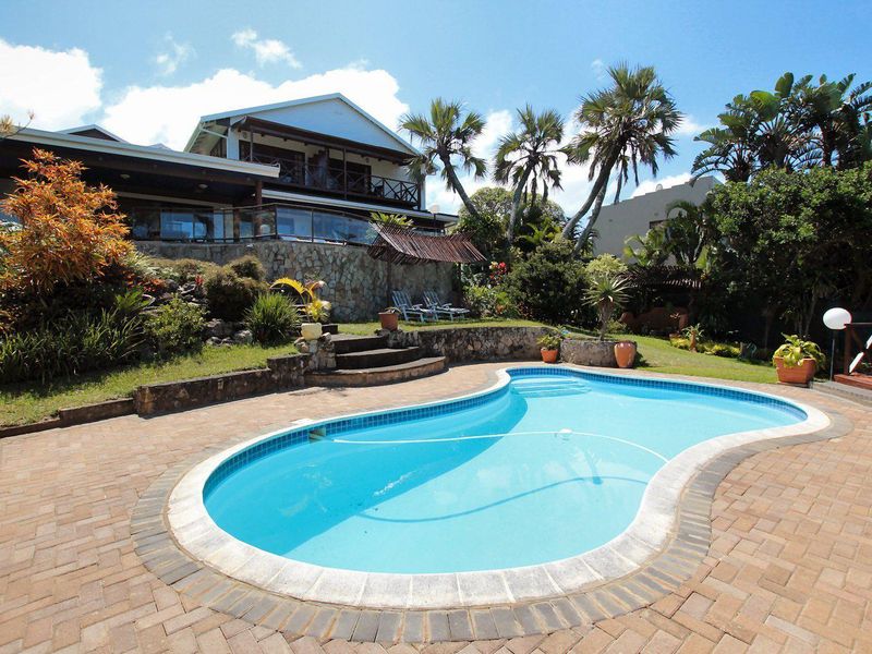 A well-maintained outdoor pool area featuring a paved deck, stone retaining walls, landscaped garden, and a view of the multi-level residential property.