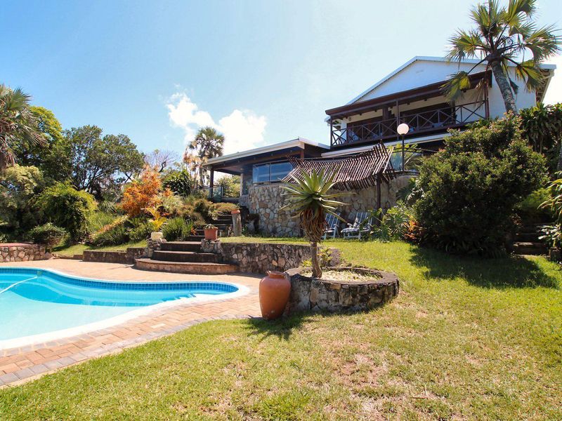 A well-maintained outdoor pool area featuring a stone-clad retaining wall, brick coping, and a spacious garden with a view of the elevated house balcony.