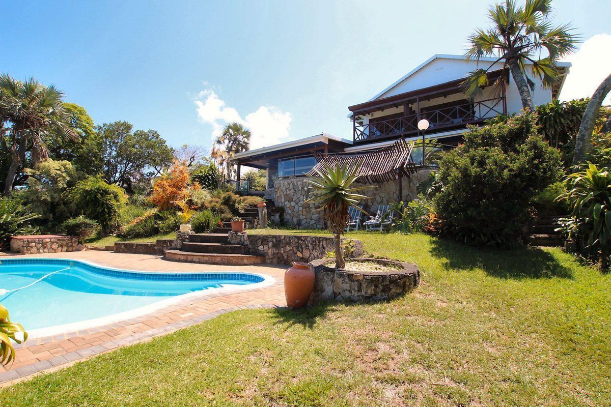 A well-maintained outdoor pool area featuring a stone-clad retaining wall, brick coping, and a spacious garden with a view of the elevated house balcony.