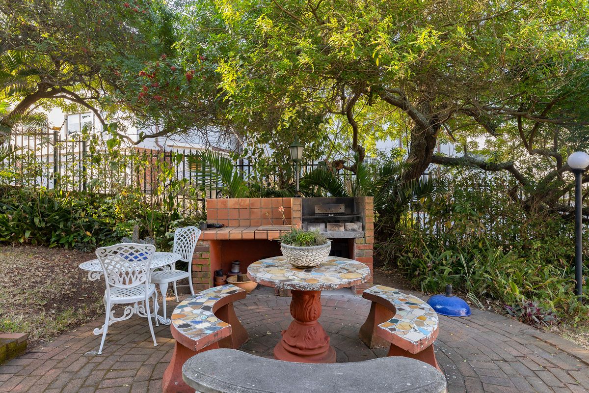 A private rear garden area featuring a built-in brick braai, paved patio space, and established trees, providing a functional outdoor entertainment setting.