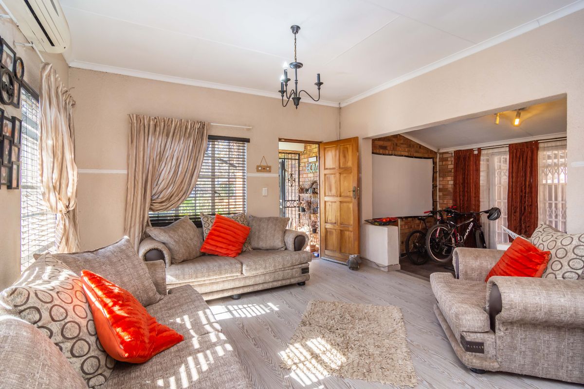 A spacious living room featuring laminate flooring, an exposed brick accent wall, and a ceiling-mounted chandelier, with an adjoining area currently used for storage.