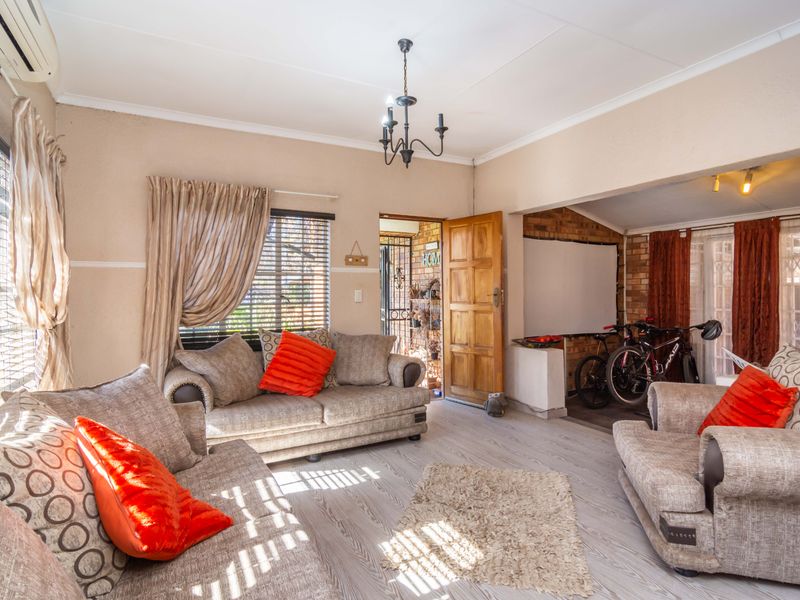 A spacious living room featuring laminate flooring, an exposed brick accent wall, and a ceiling-mounted chandelier, with an adjoining area currently used for storage.