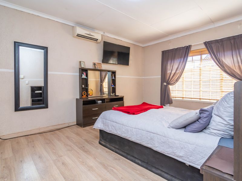 A bedroom featuring light-toned laminate flooring, a wall-mounted air conditioning unit, and window blinds, offering a functional and neutral living space.