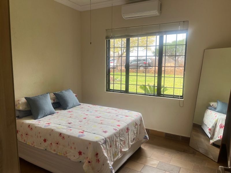 A functional bedroom featuring tiled flooring, a wall-mounted air conditioning unit, and large windows fitted with security bars and blinds overlooking the garden.