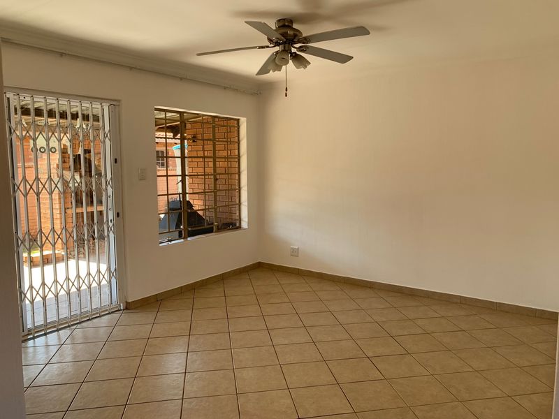 An empty living room featuring neutral tiled flooring, a ceiling fan, and secure access to the exterior via a security gate and window bars.