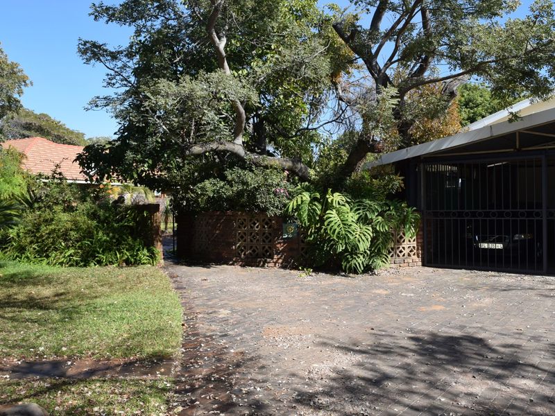Front exterior view featuring a paved driveway, a covered carport with security gate, and established garden landscaping with mature trees.