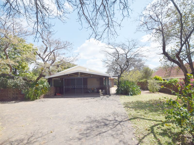 Front view of the property featuring a paved driveway, a covered carport with a security gate, and established garden surroundings.