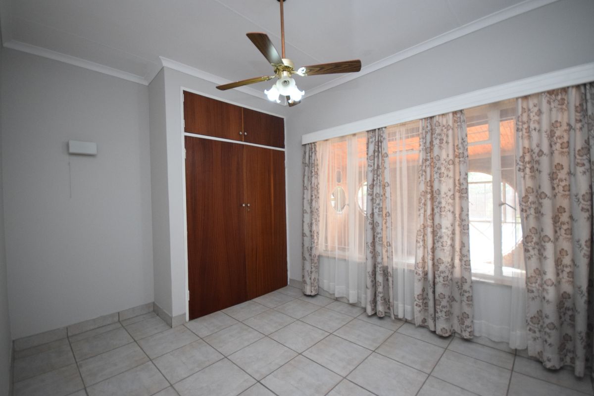 A bright, empty bedroom featuring tiled flooring, built-in wooden cupboards, and a ceiling fan, offering a clean canvas for personalization.
