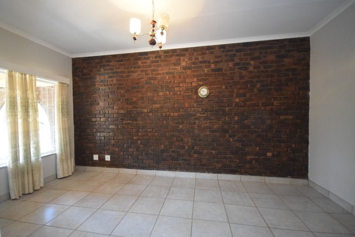 A spacious living area featuring a prominent exposed brick feature wall, neutral ceramic floor tiles, and a classic ceiling light fixture.
