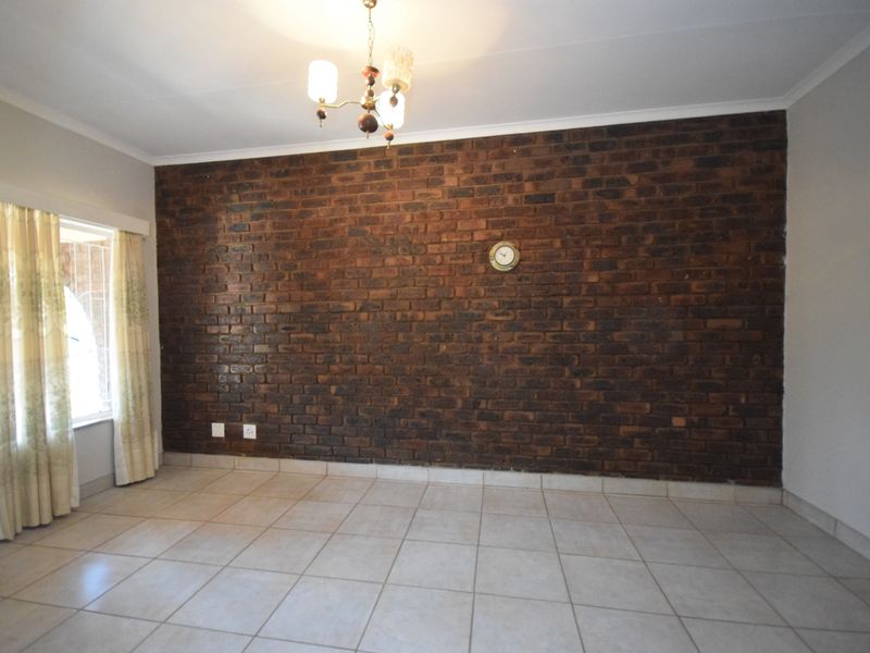 A spacious living area featuring a prominent exposed brick feature wall, neutral ceramic floor tiles, and a classic ceiling light fixture.