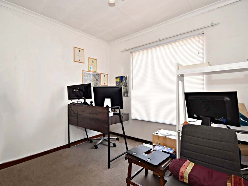 A functional home office space featuring carpeted flooring, neutral walls, and a window fitted with venetian blinds.