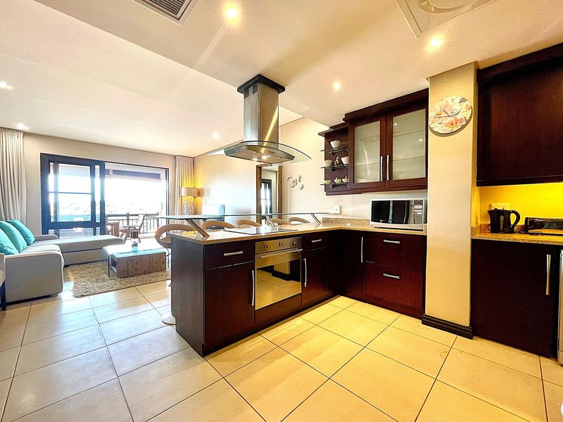 Open-plan kitchen featuring dark wood cabinetry, stone countertops, built-in oven, and stainless steel extractor fan, flowing into a bright living area with balcony access.