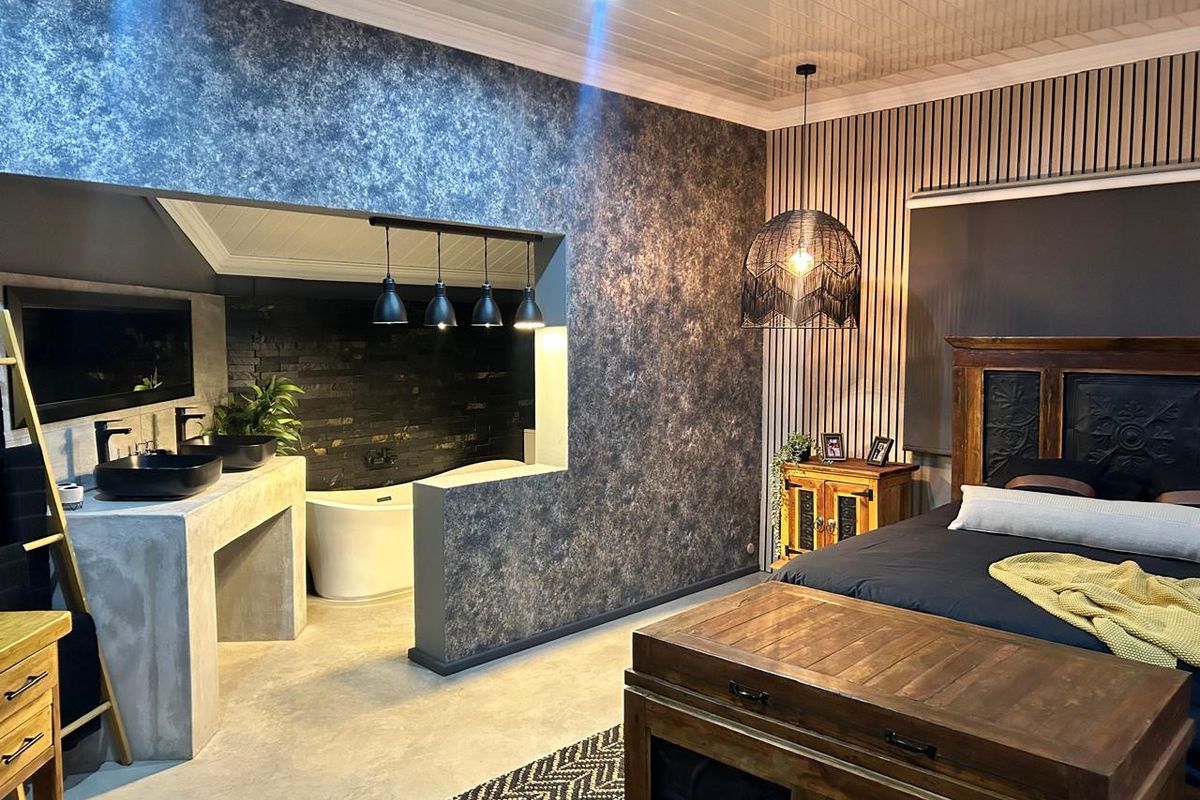 A modern bedroom featuring an open-plan ensuite with a concrete vanity, dual vessel sinks, and a freestanding bathtub against an exposed brick wall.