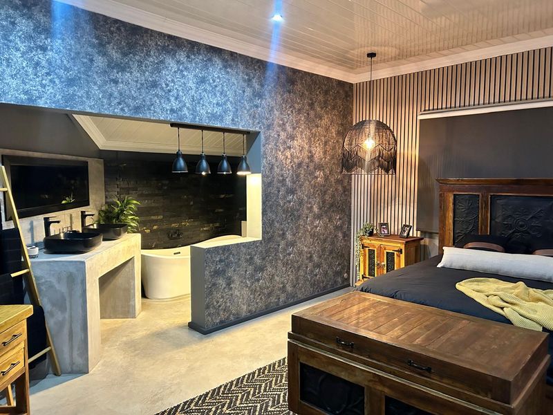 A modern bedroom featuring an open-plan ensuite with a concrete vanity, dual vessel sinks, and a freestanding bathtub against an exposed brick wall.