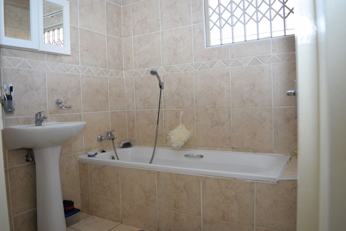 A functional bathroom featuring a pedestal basin, a bathtub with a handheld shower mixer, and neutral wall tiling with a security-fitted window.