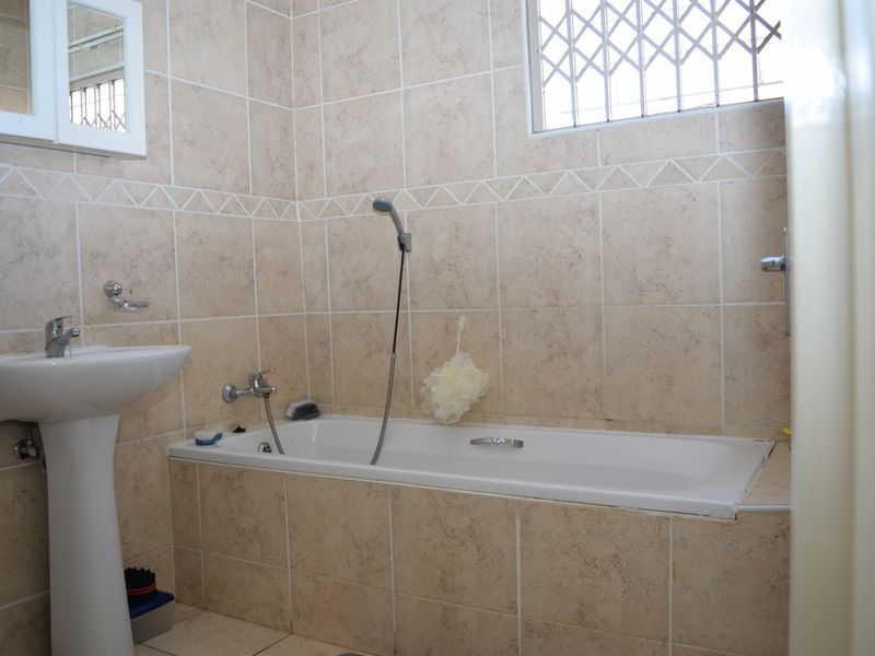 A functional bathroom featuring a pedestal basin, a bathtub with a handheld shower mixer, and neutral wall tiling with a security-fitted window.