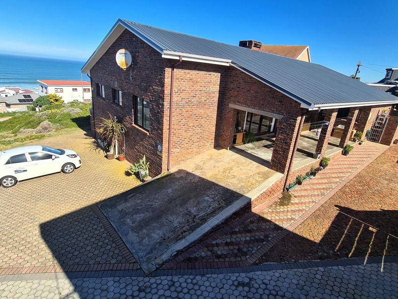 An elevated view of a brick-faced house featuring a metal roof, paved driveway, and a covered patio area with ocean views.