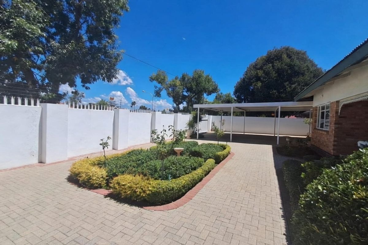 A well-maintained rear exterior area featuring a paved driveway, a covered carport, and a neat landscaped garden bed enclosed by a secure perimeter wall.