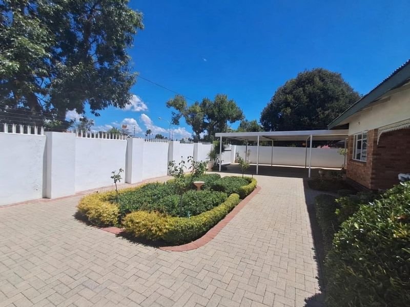 A well-maintained rear exterior area featuring a paved driveway, a covered carport, and a neat landscaped garden bed enclosed by a secure perimeter wall.
