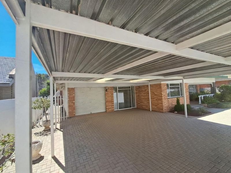 A front exterior view featuring a paved driveway, a covered carport with a corrugated metal roof, and a brick facade with a roller shutter garage door.