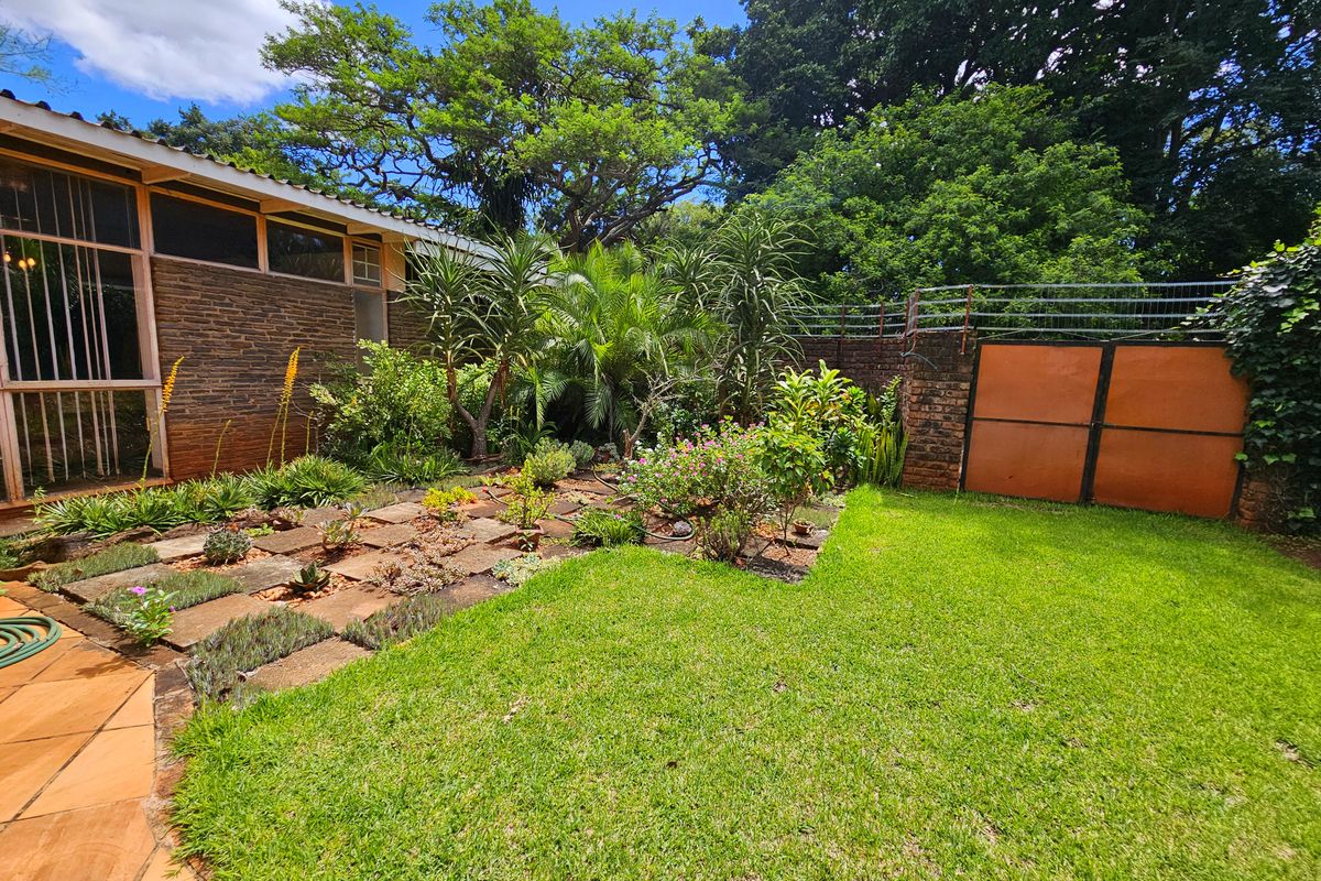 A well-maintained rear garden featuring a paved patio area, established landscaping, and a secure perimeter wall with a metal gate.