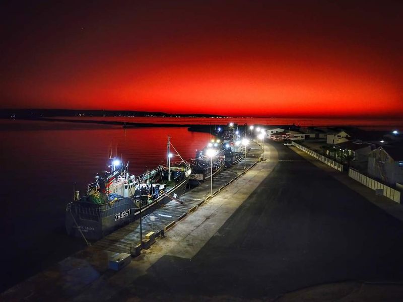 Aerial view of a harbour at sunset, featuring a jetty with docked fishing vessels and illuminated street lighting along the waterfront.