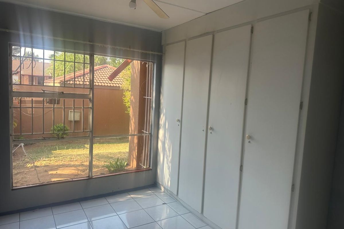 A bright, empty bedroom featuring tiled flooring, built-in cupboards, and a ceiling fan, with large windows overlooking the rear garden area.