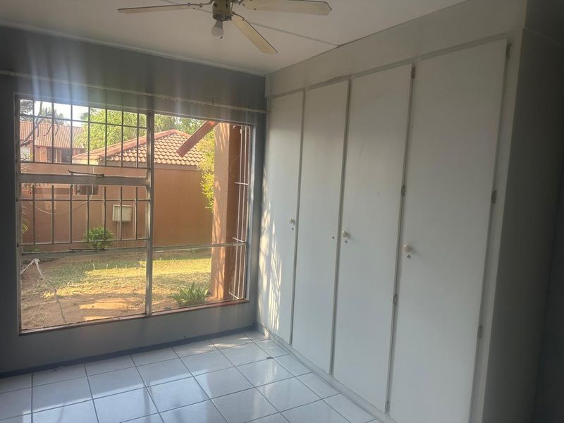 A bright, empty bedroom featuring tiled flooring, built-in cupboards, and a ceiling fan, with large windows overlooking the rear garden area.