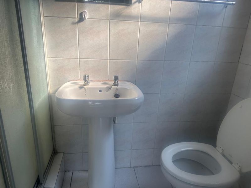 A functional bathroom featuring a pedestal basin, toilet, and shower enclosure with tiled walls and flooring, requiring cosmetic updates to improve its overall condition.