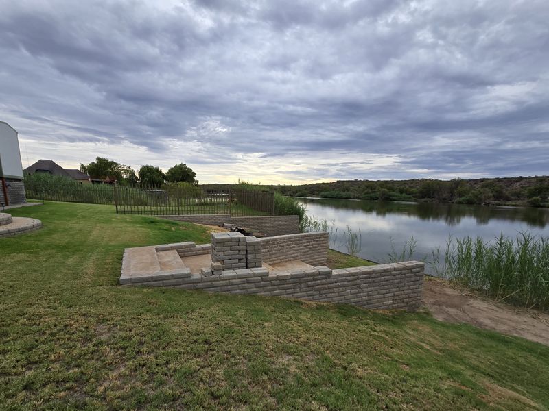 Rear garden area featuring a riverfront view, lawn, and a partially constructed brick fire pit and retaining wall structure.
