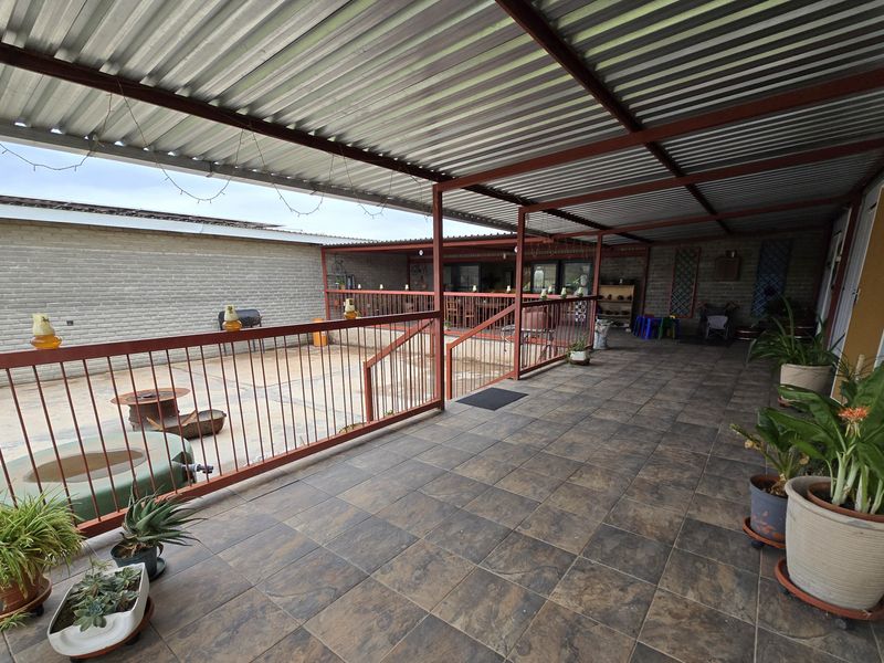 A spacious covered patio area featuring tiled flooring, a corrugated metal roof, and metal safety railings, overlooking a paved outdoor space.
