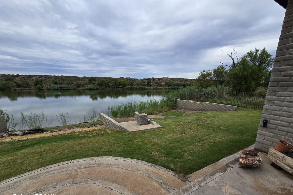 Rear garden area featuring a paved patio, built-in brick braai, and retaining walls, overlooking a scenic riverfront landscape.