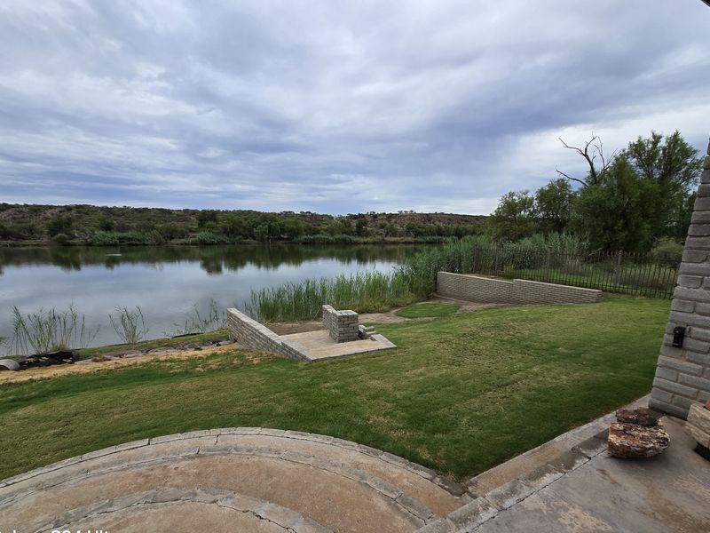 Rear garden area featuring a paved patio, built-in brick braai, and retaining walls, overlooking a scenic riverfront landscape.